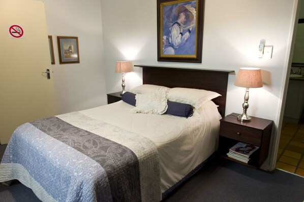 A neatly made bed with a white and gray patterned bedspread, two pillows, and two bedside lamps. There is a framed picture on the wall above the bed and a no-smoking sign on the door. - Primavera Guest House (Bloemfontein)
