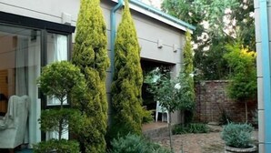 A courtyard with tall, narrow trees, a brick pathway, and a white chair. - Primavera Guest House (Bloemfontein)