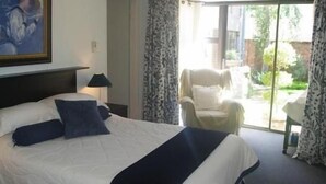 A hotel room with a large bed, a nightstand with a lamp, a chair, and a view of the garden through a sliding door. - Primavera Guest House (Bloemfontein)