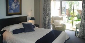 Habitación de hotel con una cama grande, una mesita de noche con lámpara, una silla y vistas al jardín a través de una puerta corredera.