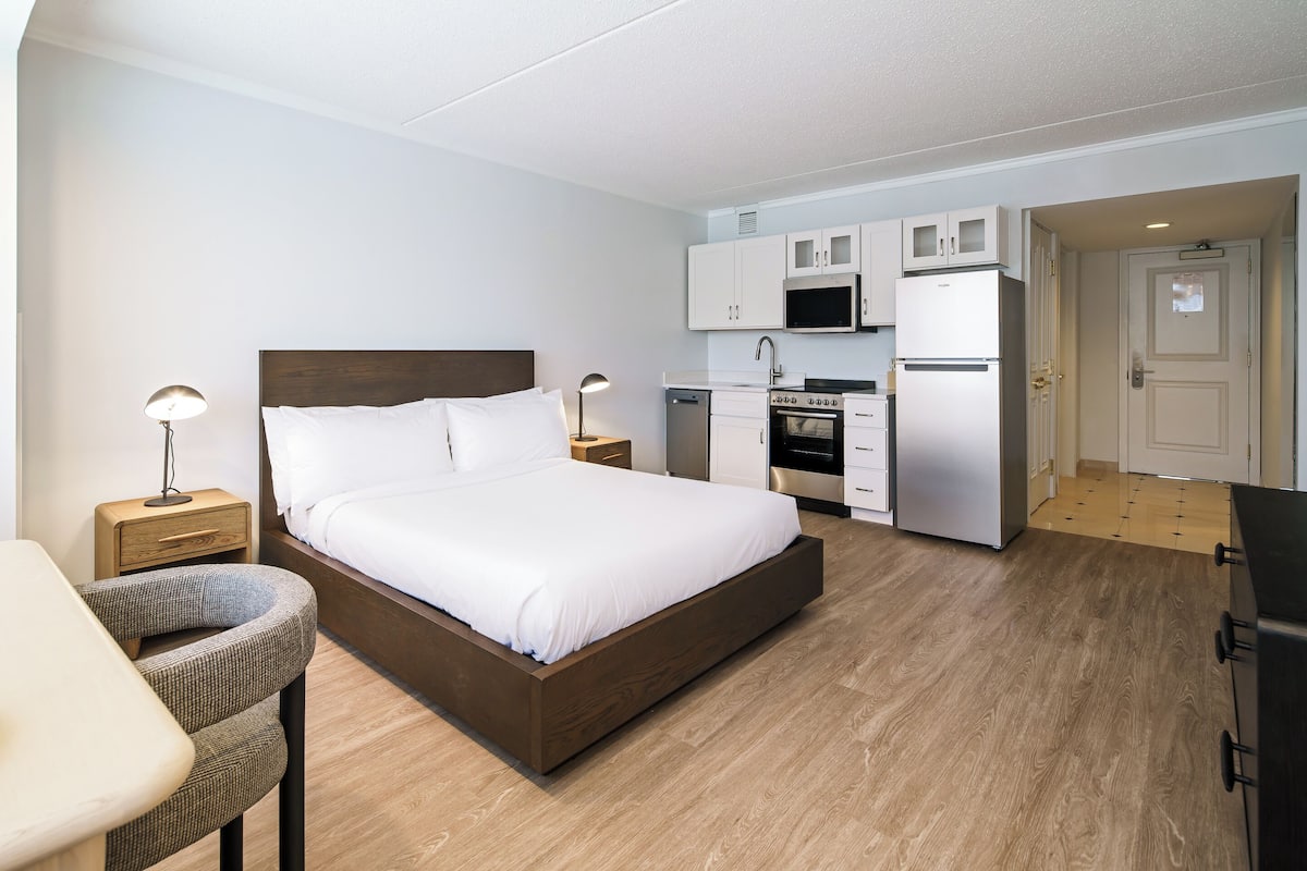 A bedroom with a bed, bedside tables, a chair, a desk, and a kitchenette with a microwave, stove, and refrigerator.