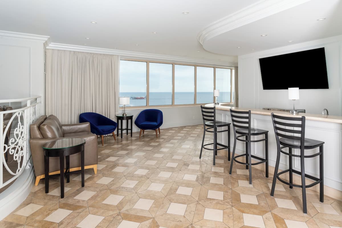 A modern hotel room with a large window, a flat-screen TV, a bar area, and a seating area with a sofa and chairs.