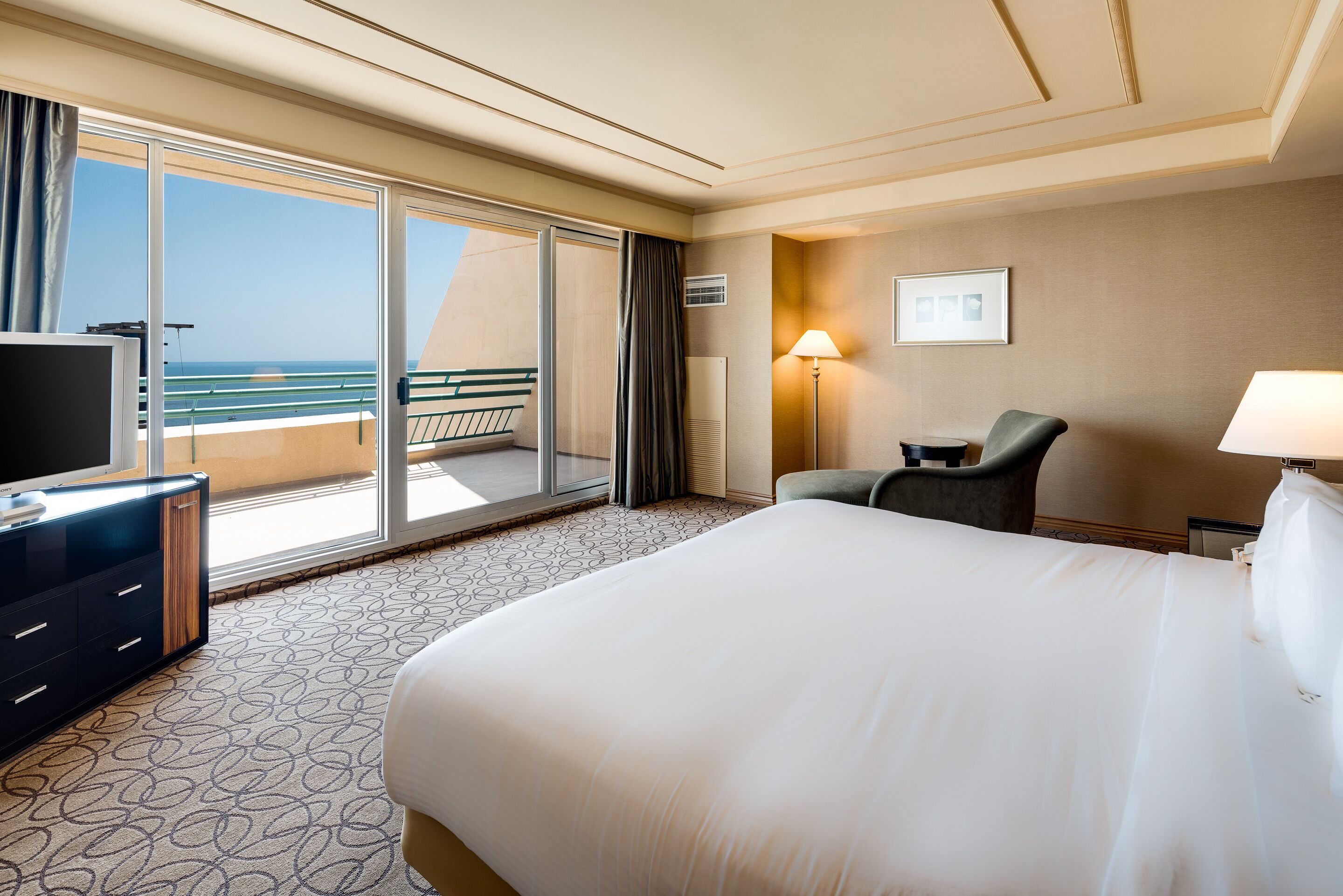 Habitación de hotel con una cama grande, televisión, escritorio y una silla. Tiene un balcón con vistas al océano.