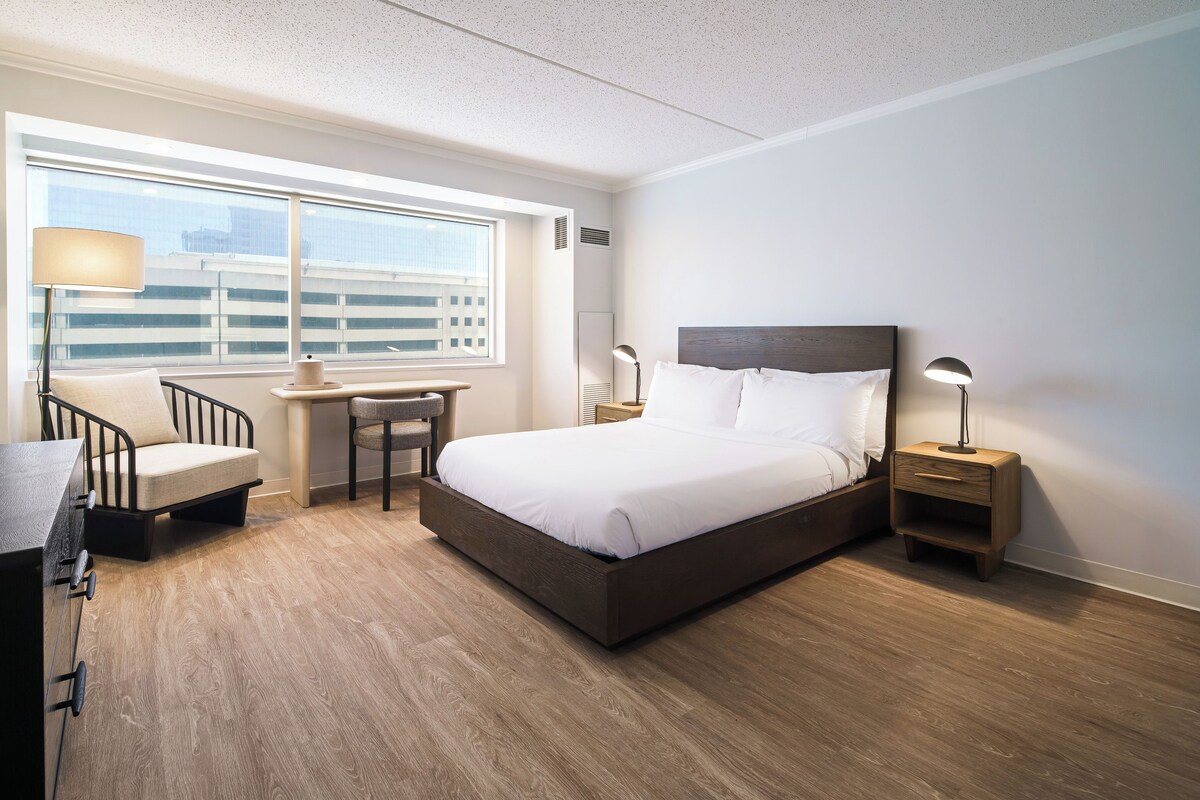 A hotel room with a bed, desk, chair, lamp, and a window with a city view.