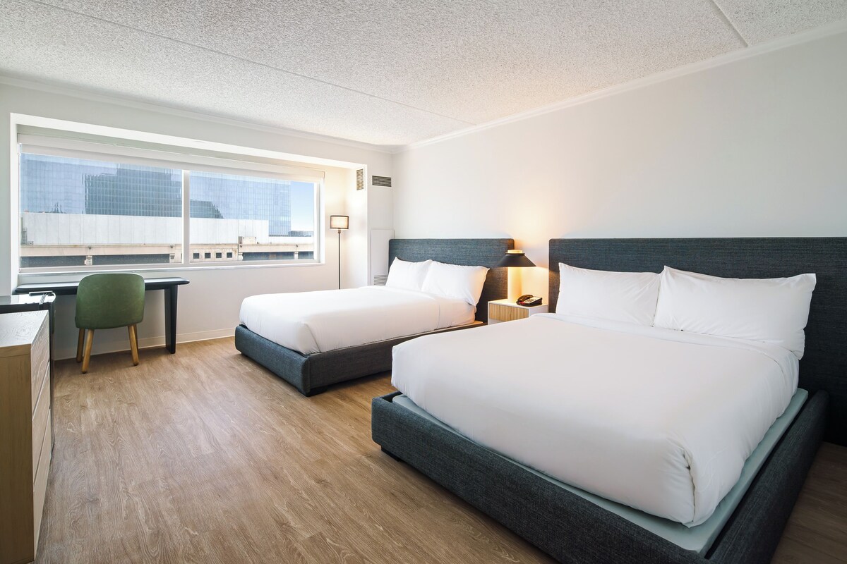A hotel room with two beds, a desk, a chair, and a window with a city view.
