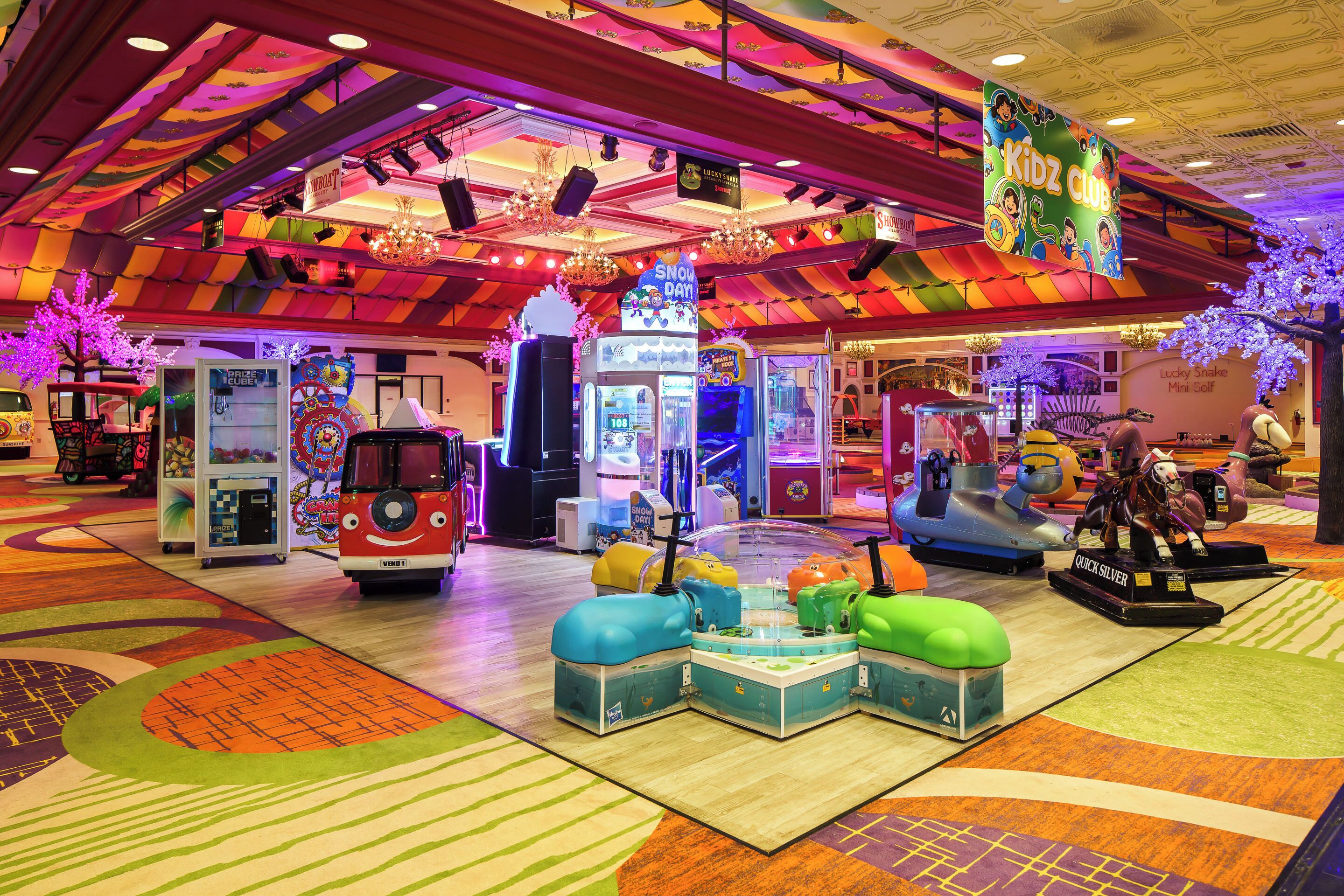 A vibrant indoor play area with arcade games, a fire truck ride-on, and a 'Kidz Club' sign.