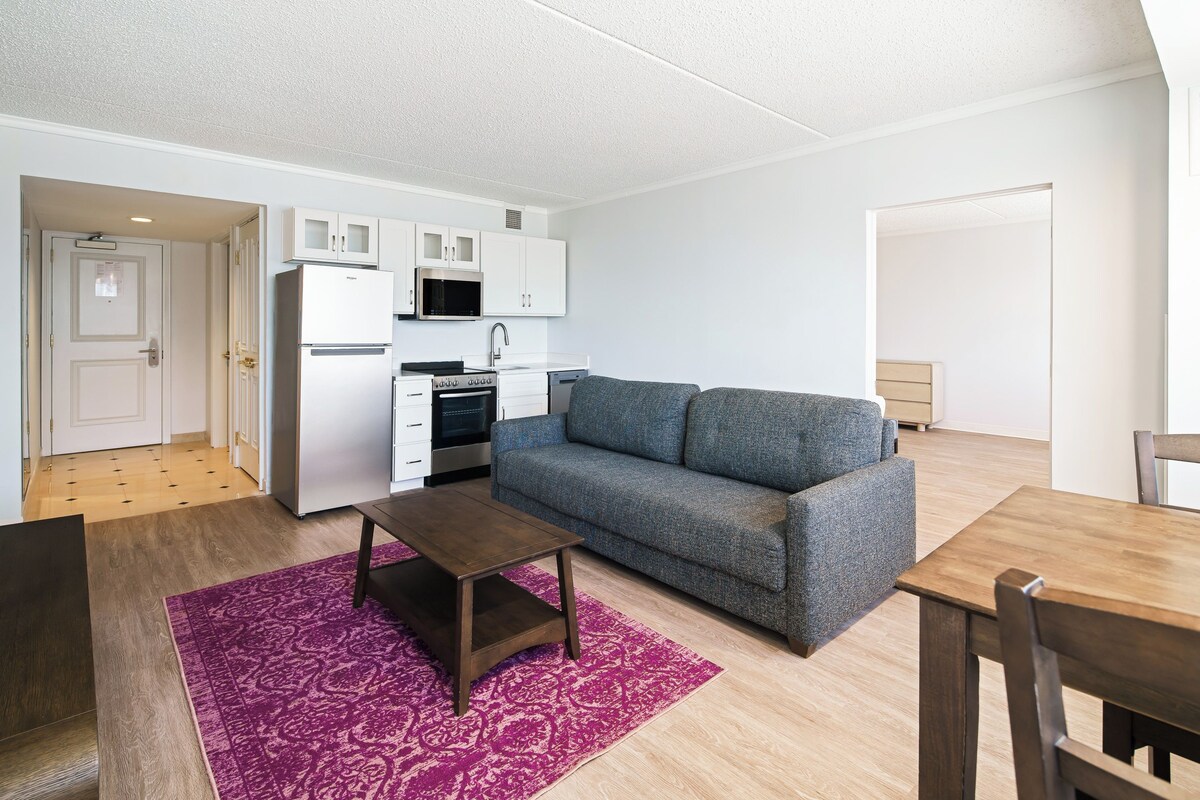 A compact living space with a kitchenette, a gray sofa, a wooden table, and a purple rug.