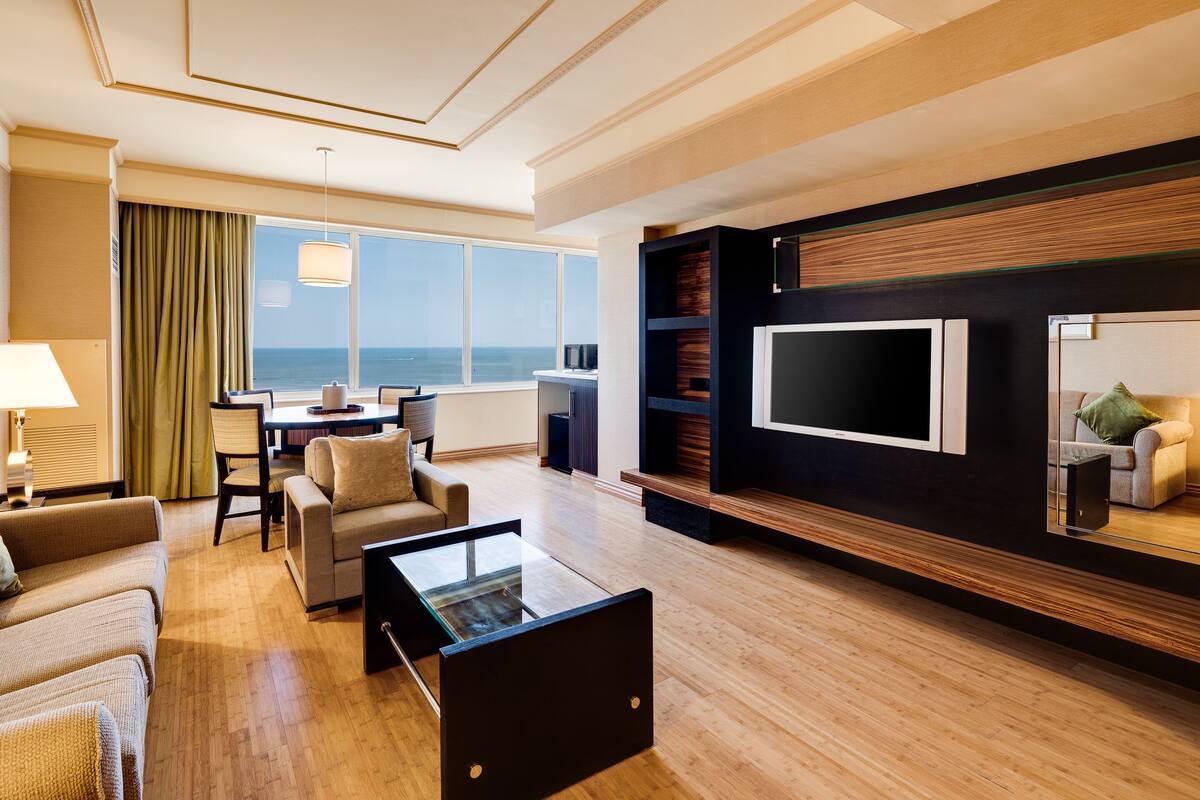 A modern hotel room with a large window, a flat-screen TV, a sofa, a coffee table, and a dining area.