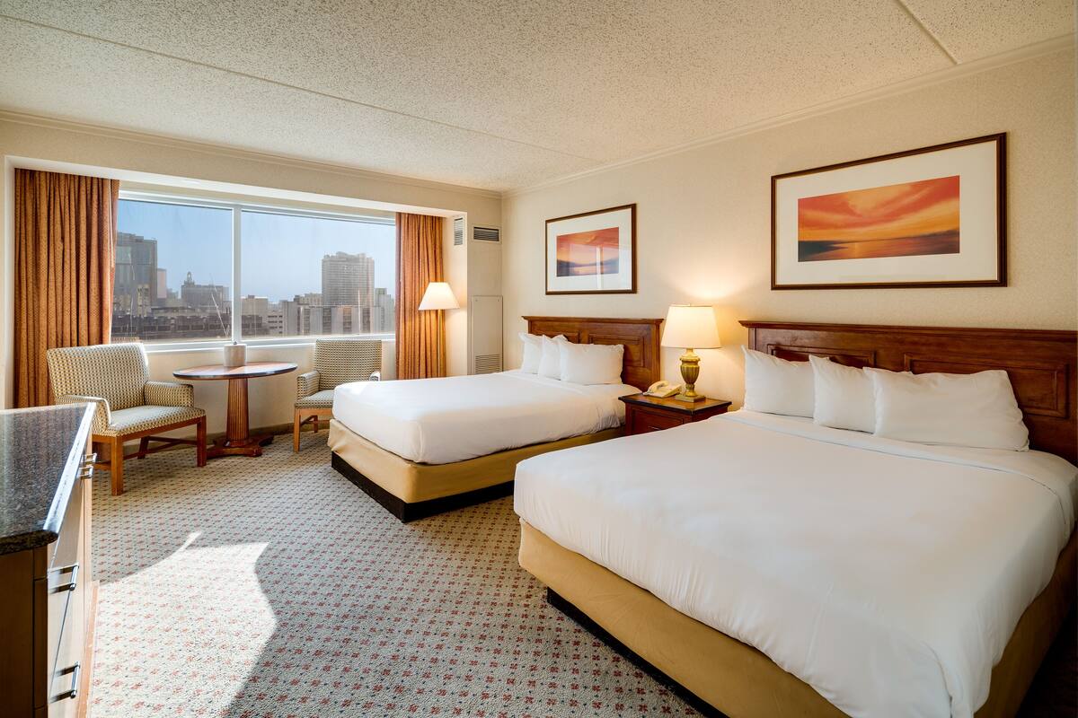 A hotel room with two beds, a desk, a chair, a small table, and a window with a city view.