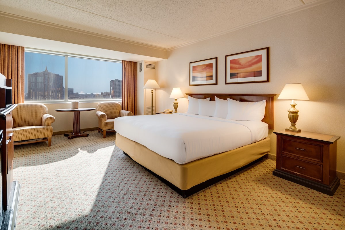 A hotel room with a large bed, two bedside tables with lamps, a small table with a chair, and a view of the city through large windows.