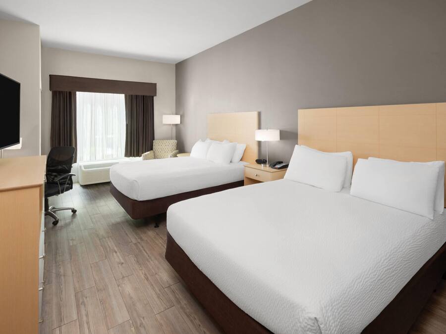 Habitación de hotel con dos camas, un escritorio con televisor, una silla, una ventana con cortinas y una mesita de noche con lámpara.