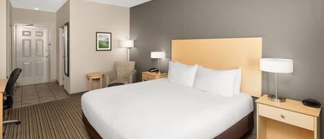 A hotel room with a large bed, a desk, a chair, a nightstand with a lamp, and a picture on the wall.