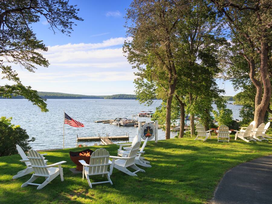 Terraza junto al lago con sillas Adirondack blancas, un brasero y una bandera estadounidense.