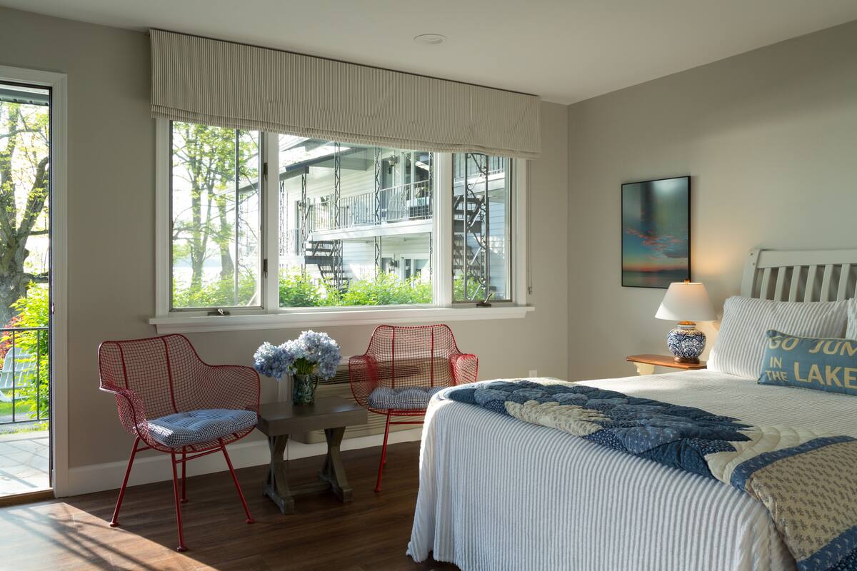 Ein Schlafzimmer mit einem großen Fenster, einem Bett mit einer blau-weißen Steudecke, zwei roten Sesseln, einem kleinen Tisch mit Blumenvase und einem Nachttisch mit Lampe.