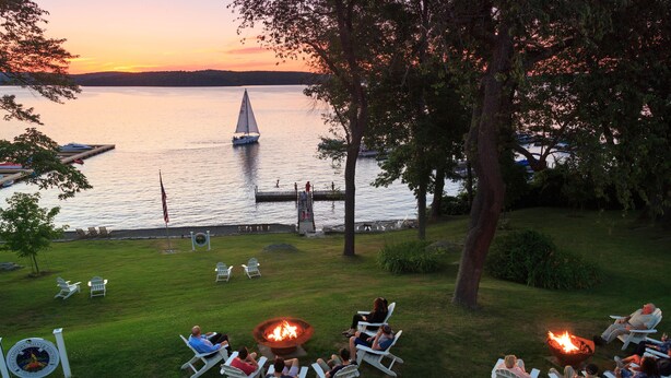 A lakeside patio with lounge chairs arranged around a central fire pit, overlooking a calm lake with a sailboat in the distance.