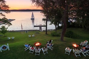 A lakeside patio with lounge chairs arranged around a central fire pit, overlooking a calm lake with a sailboat in the distance.