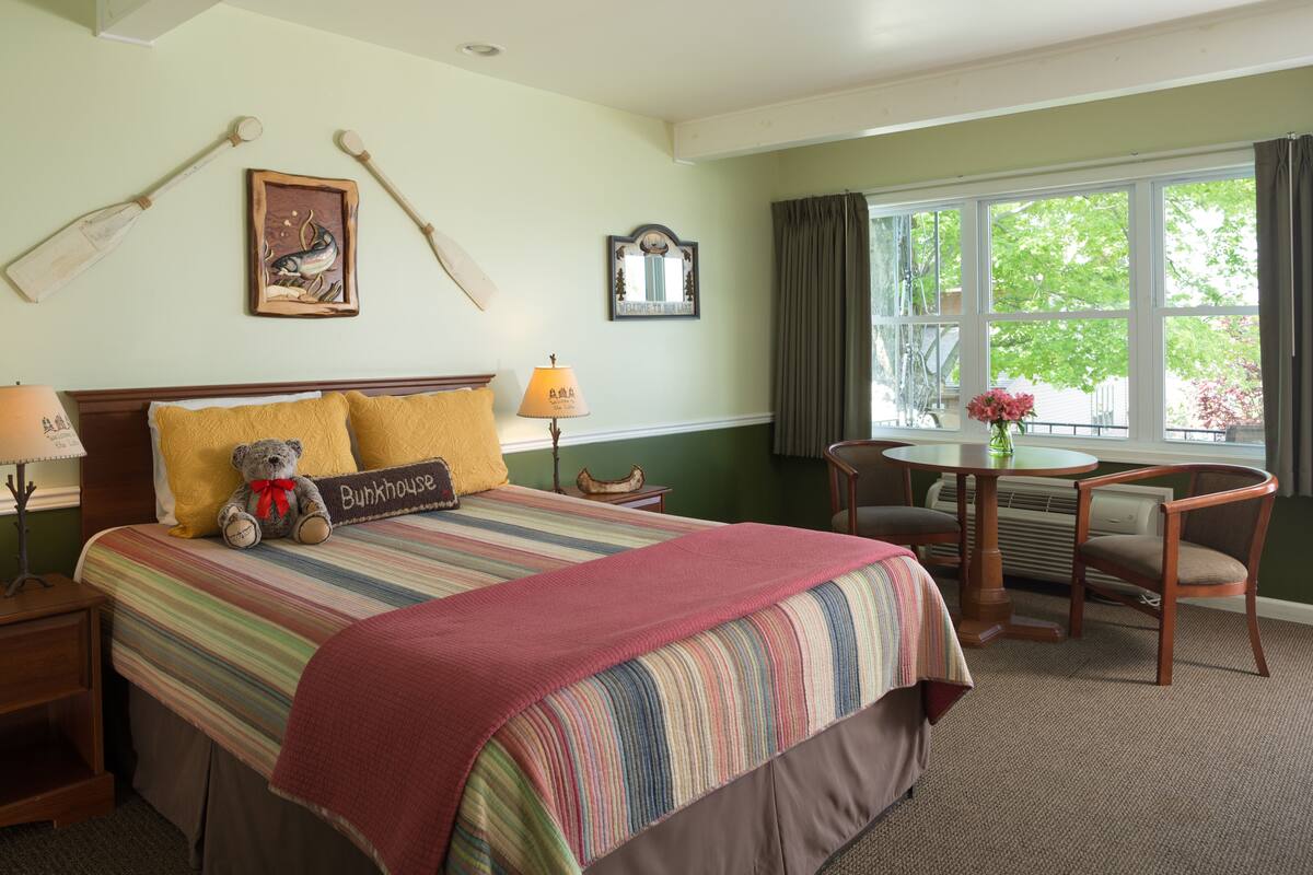Ein Hotelzimmer mit einem großen Bett, einem kleinen Tisch und einem Stuhl. Auf dem Bett liegt ein Teddybär und ein Schild mit der Aufschrift 'Bunkhouse'.