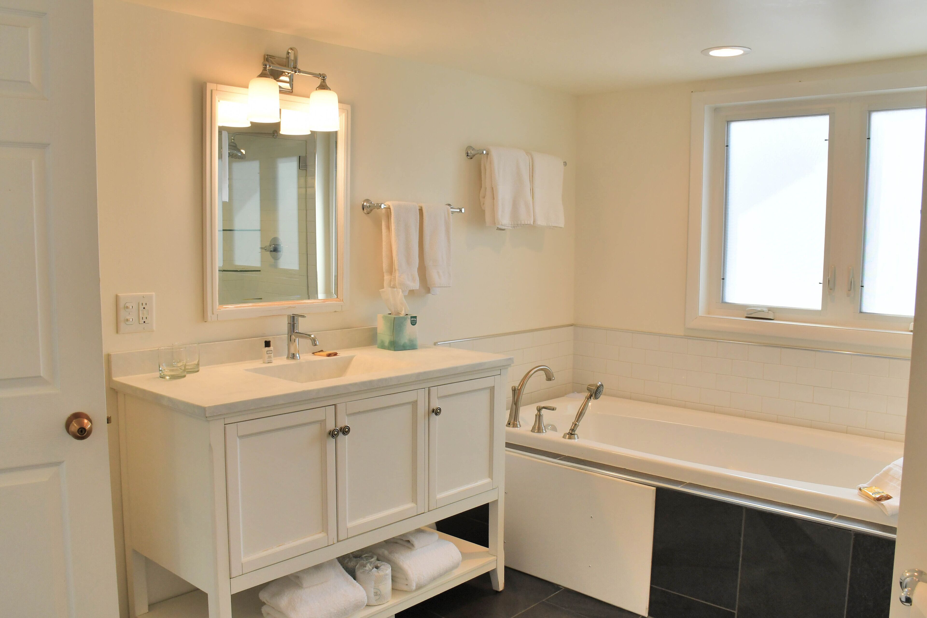 A modern bathroom with a white vanity, a large mirror, a bathtub, and a window.