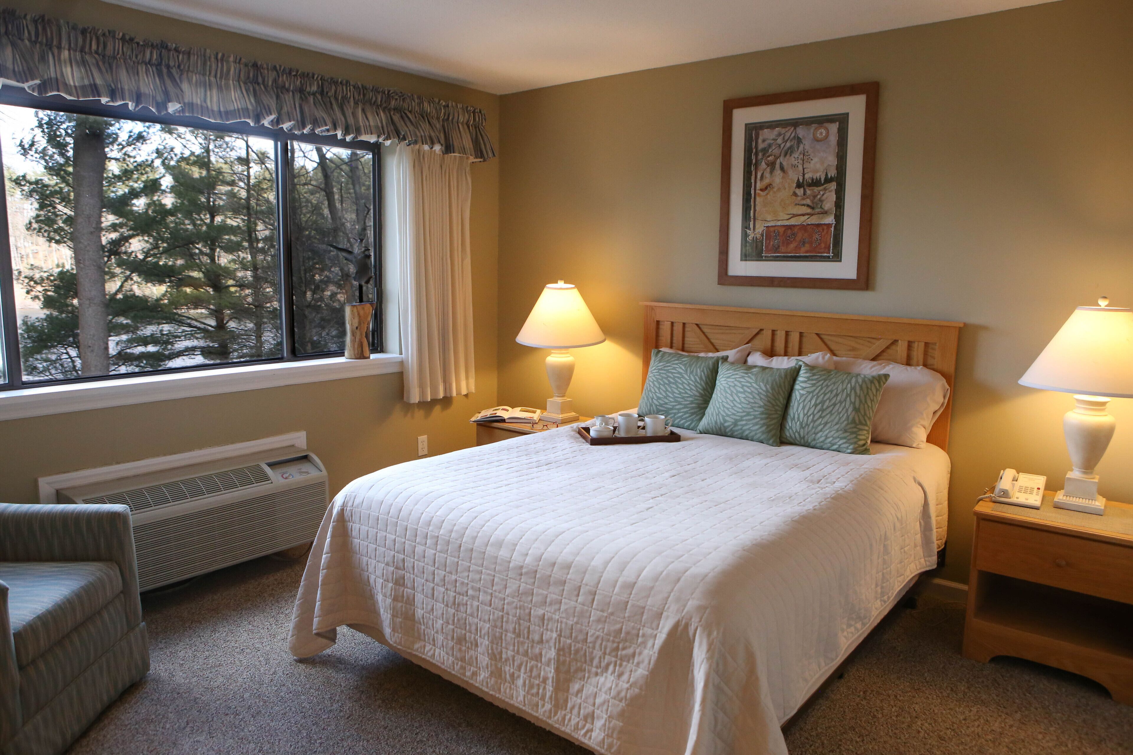 A hotel room with a large bed, two bedside tables with lamps, a chair, a window with curtains, a radiator, and a painting on the wall.