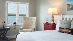 A bedroom with a large bed, a chair, a nightstand with a lamp, a window with a view of the lake, and a painting on the wall.