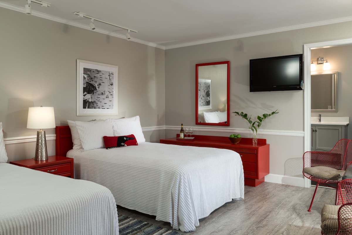 Ein Hotelzimmer mit zwei Betten, einem roten Schminktisch, einem Fernseher und einem Badezimmer mit Spiegel und Waschbecken.