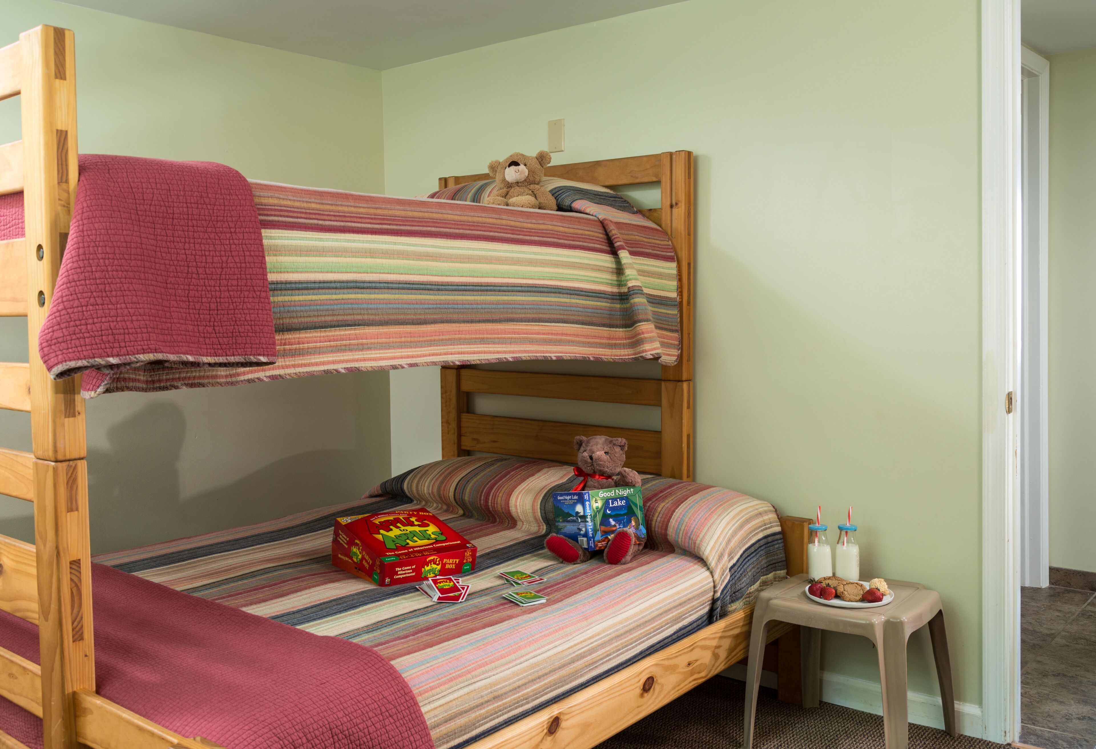 Una cama litera con una colcha a rayas, un osito de peluche, una caja de juegos y una mesita con galletas y bebidas.
