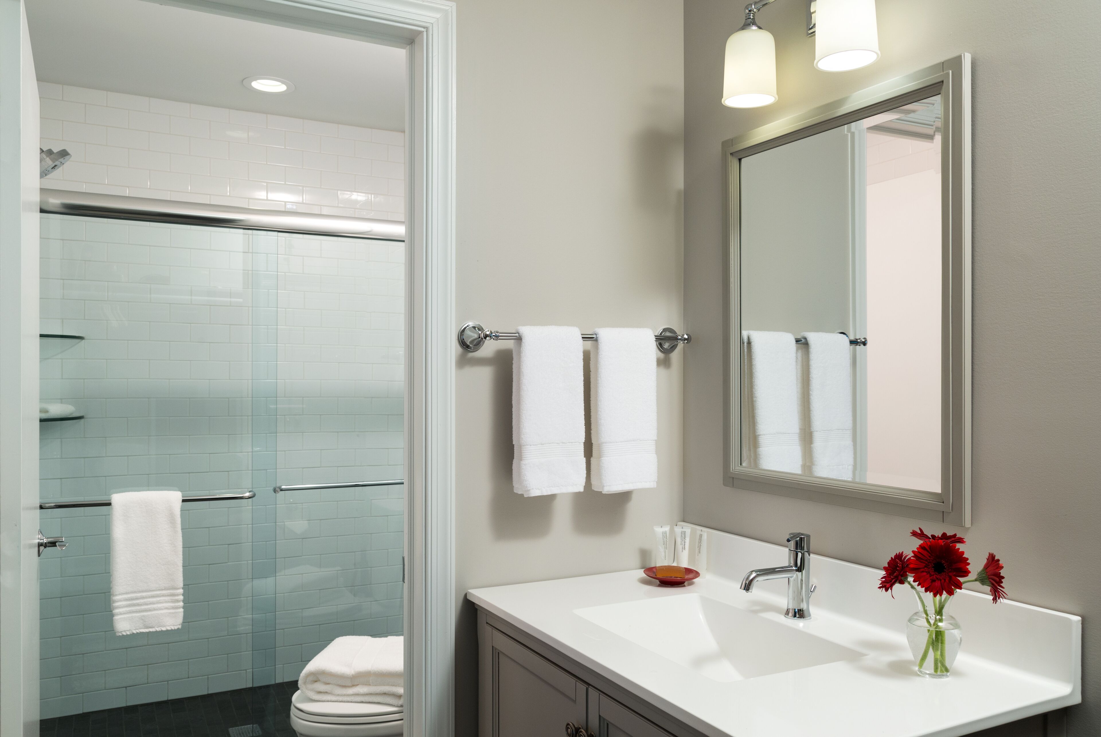 Une salle de bain moderne avec une douche à l’italienne en verre, un lavabo blanc avec un miroir au-dessus, et un vase avec des fleurs rouges posé sur le lavabo.