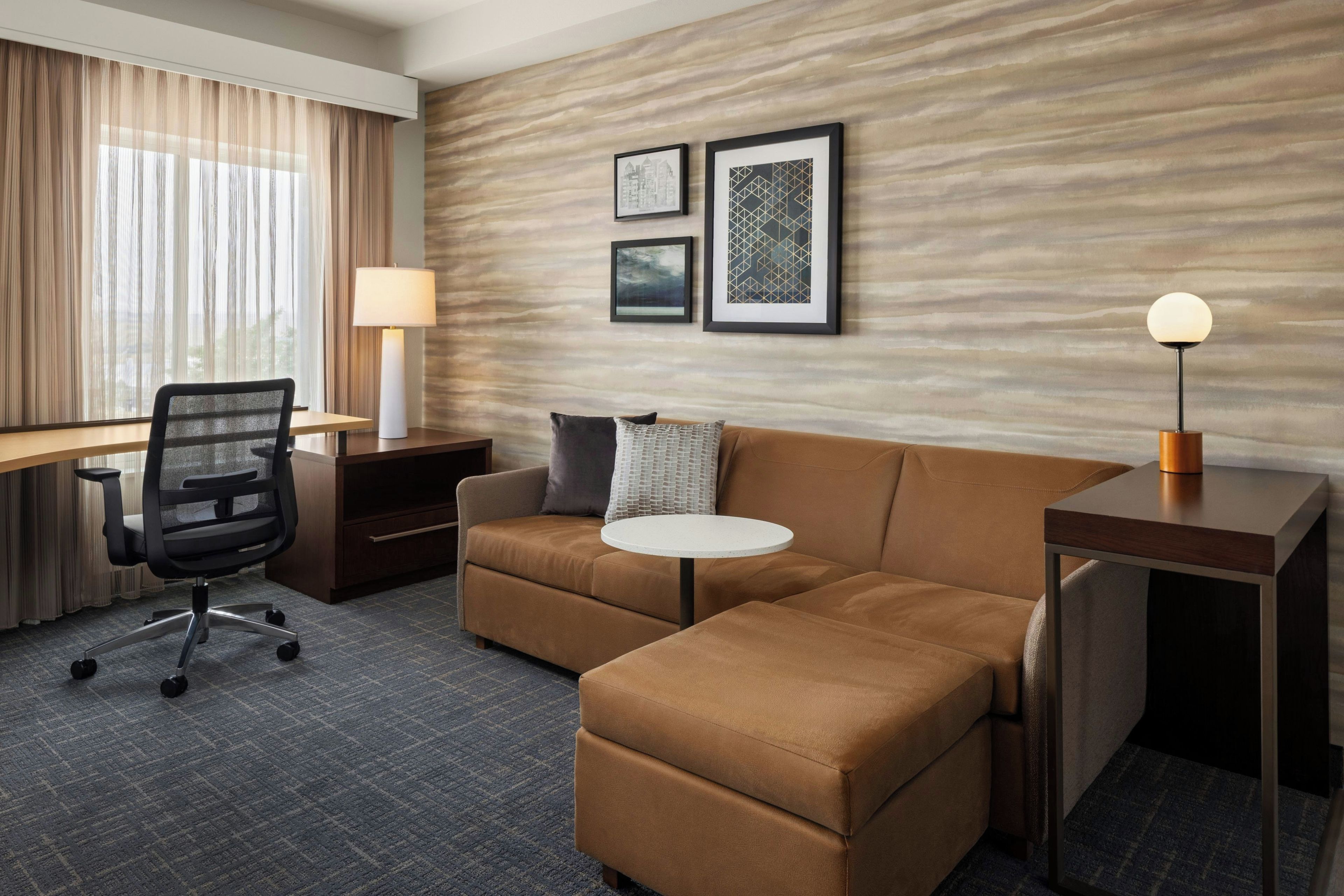 A hotel room with a sectional sofa, a desk with a chair, a small round table, and a lamp.