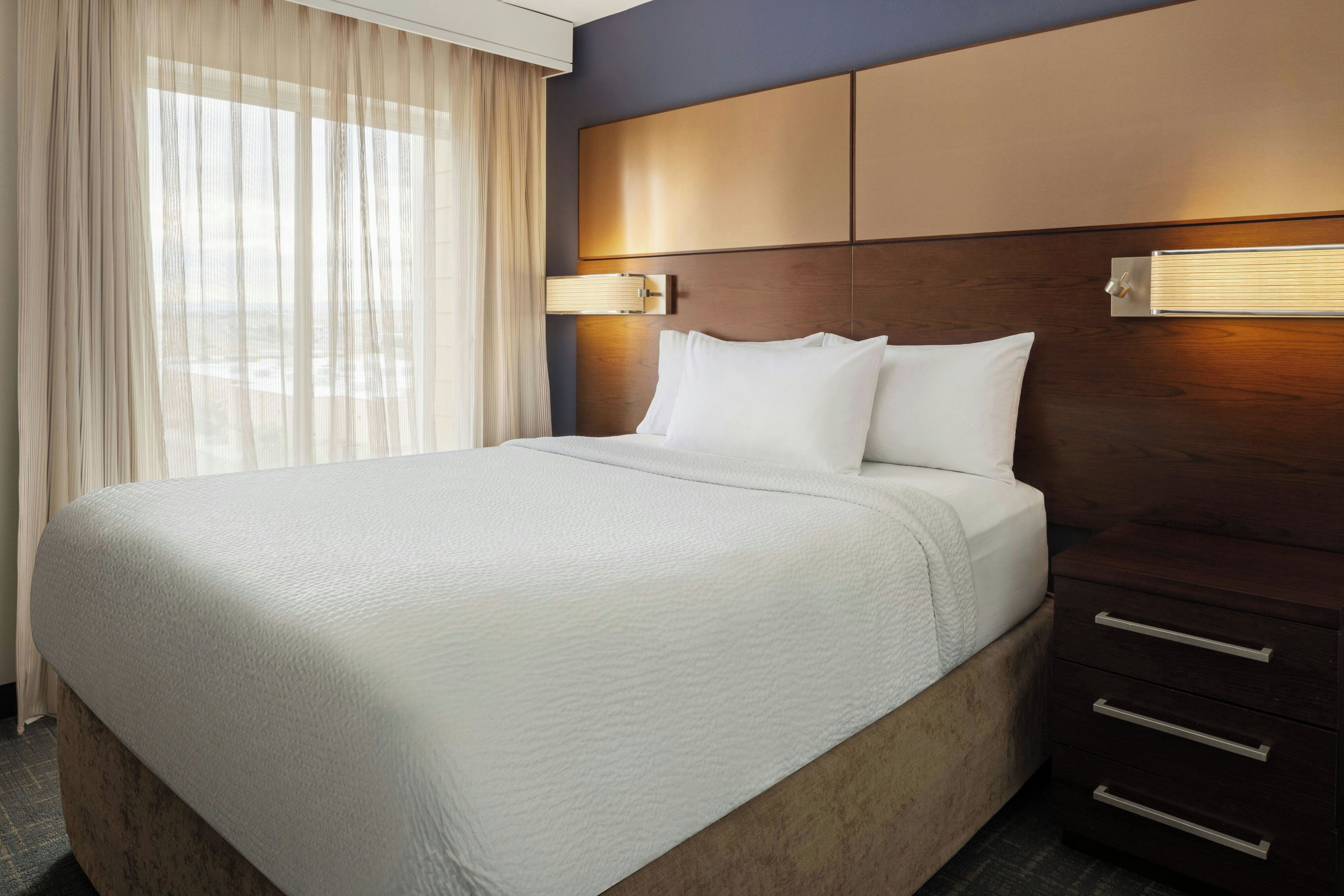 A hotel room with a large bed, a wooden headboard, a nightstand, and a window with sheer curtains.