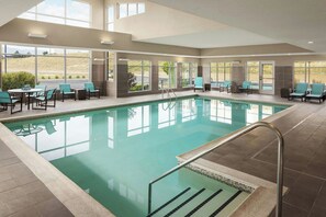 Una piscina interior con paredes de vidrio transparente, suelo de baldosas y una zona de estar con sillas y una mesa.
