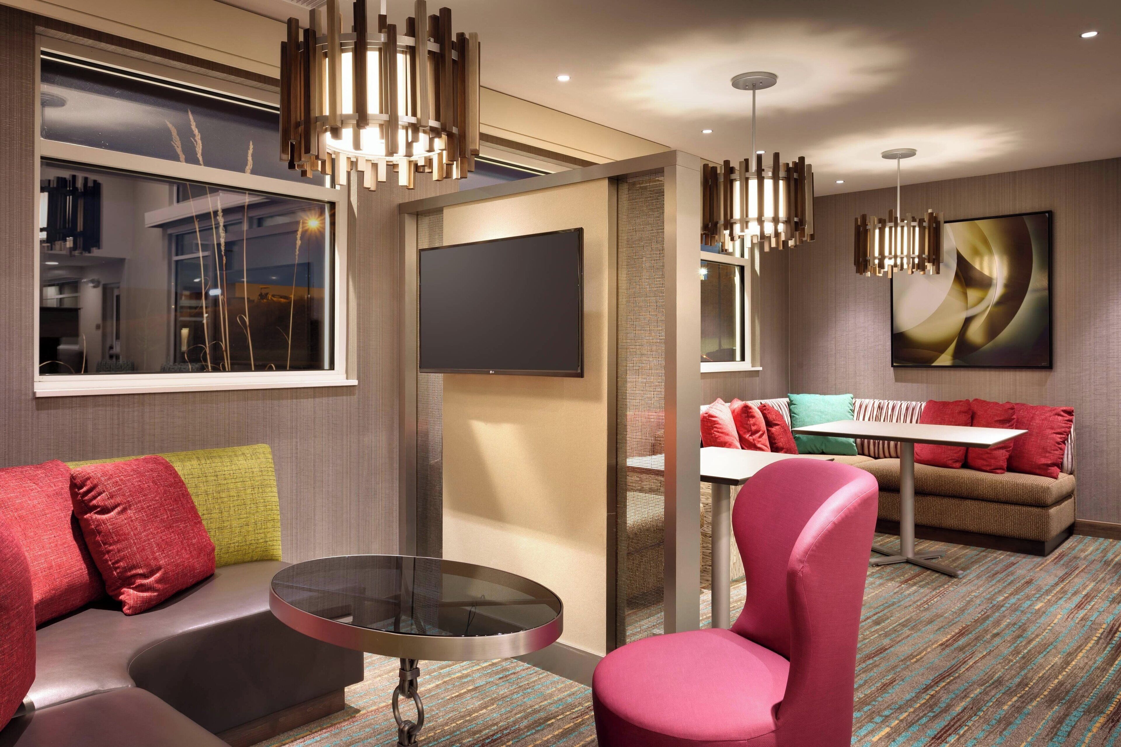 A modern hotel room with a sofa, a round table, and a pink chair. There is a flat-screen TV mounted on the wall and a painting above the sofa.