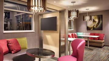 A modern hotel room with a sofa, a round table, and a pink chair. There is a flat-screen TV mounted on the wall and a painting above the sofa.