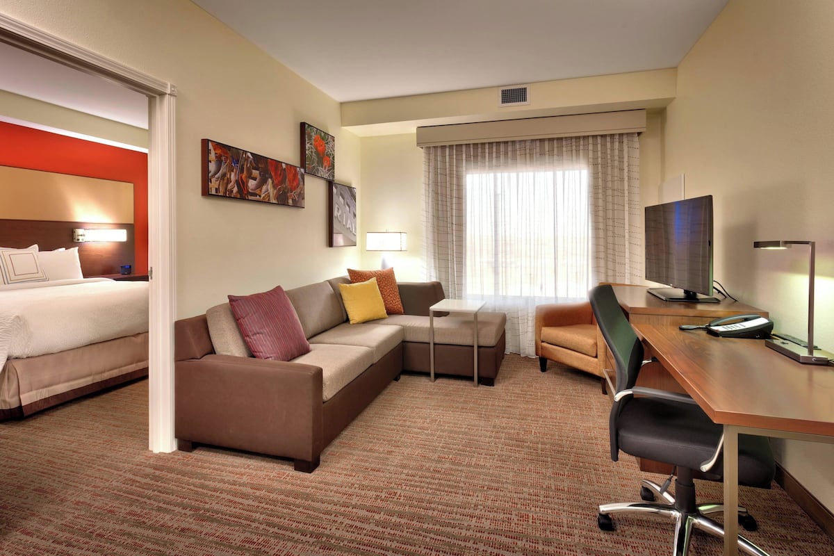 A hotel room with a bed, a sofa, a desk, and a chair. There is a window with curtains, a lamp, and a television.