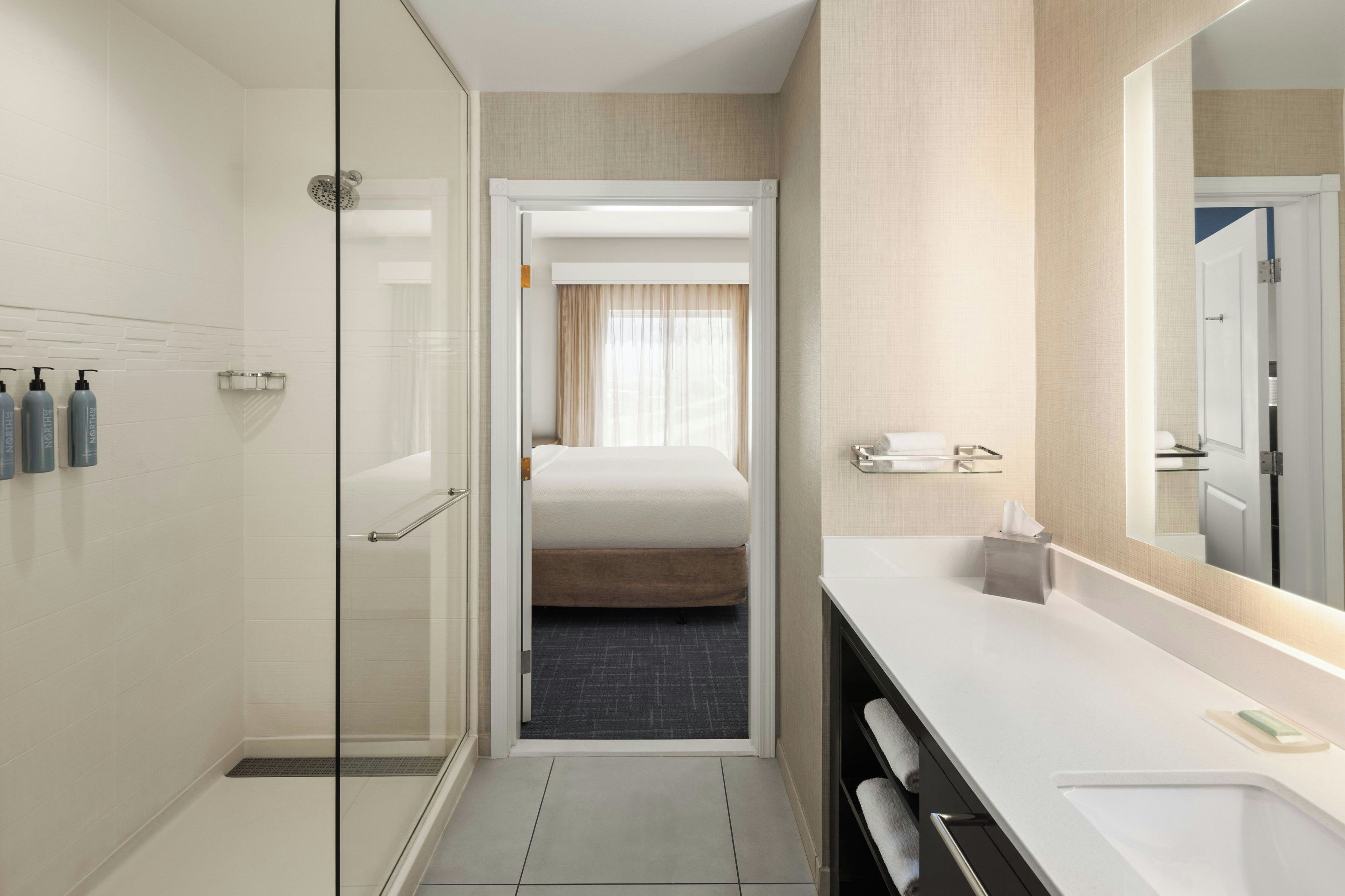 A hotel bathroom with a shower, a bed, a sink, and a mirror.