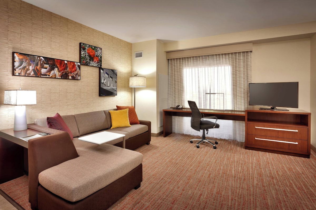 A hotel room with a sofa, a desk, a chair, a TV, and artwork on the wall.