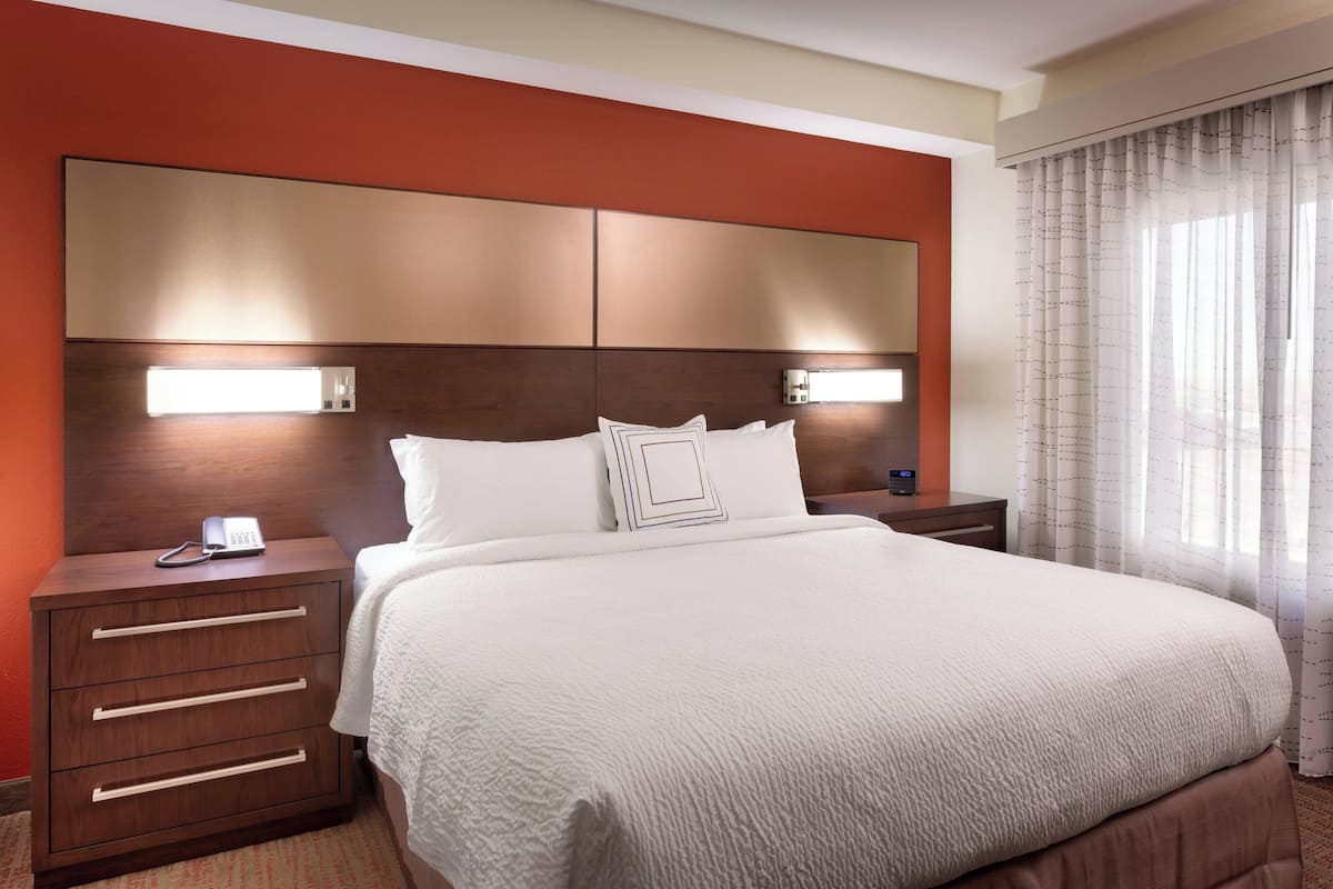 A hotel room with a large bed, a wooden nightstand with drawers, a bedside table with a phone, and a headboard with integrated lighting.