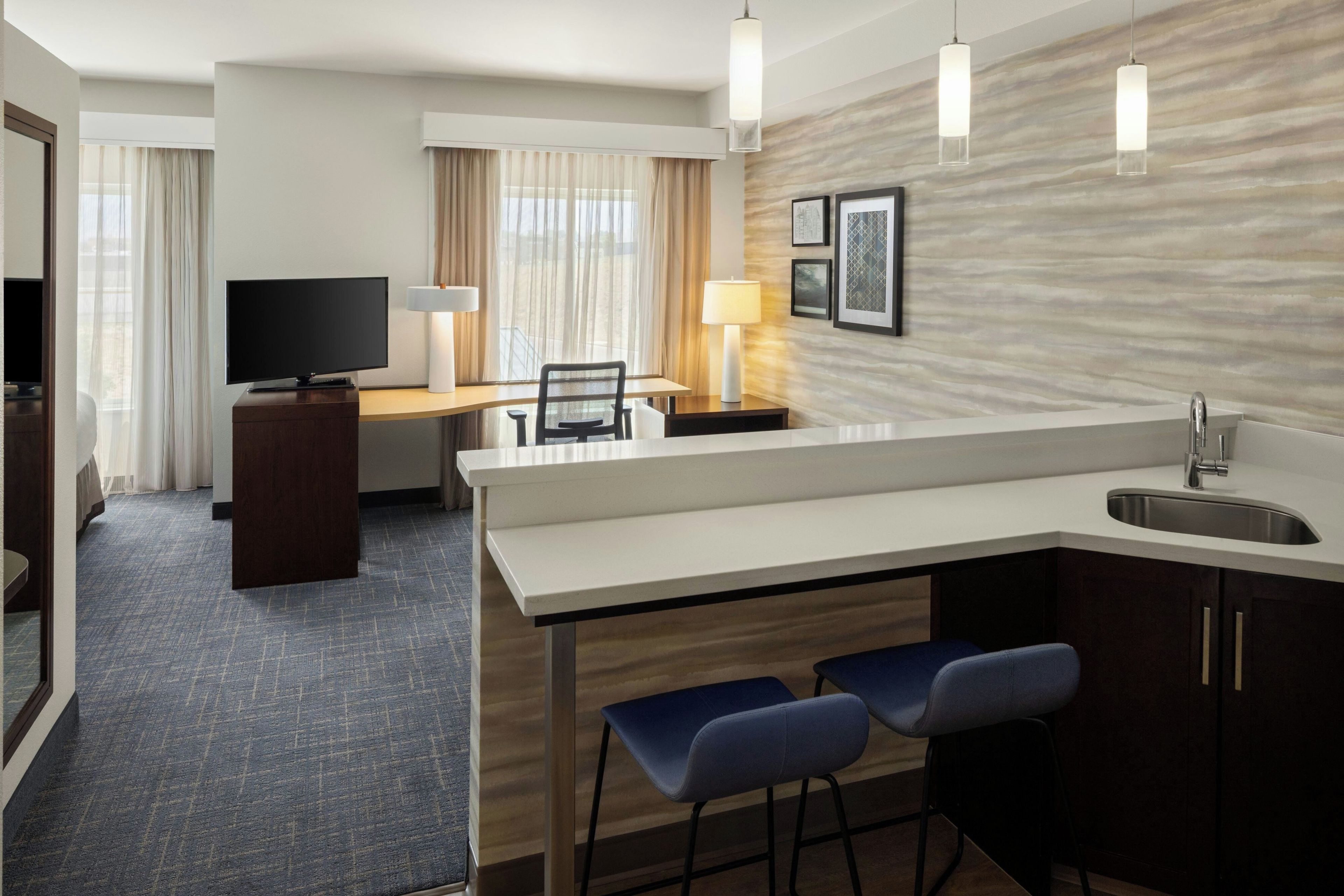 A hotel room with a kitchenette, a television, a desk, and a seating area with two chairs.