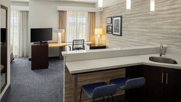 A hotel room with a kitchenette, a television, a desk, and a seating area with two chairs.