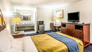 Habitación de hotel con cama, escritorio con silla, televisión, refrigerador y baño con espejo y toallero.
