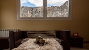 A room with a large window overlooking rocky terrain, a brown leather sofa, and a fluffy rug. - Vertical Lodge (El Chalten)