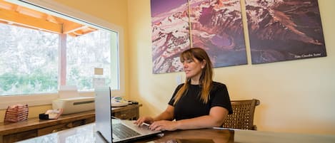 Une femme travaille à un bureau, devant un ordinateur portable. Une fenêtre et des tableaux représentant des paysages de montagne ornent le mur.
