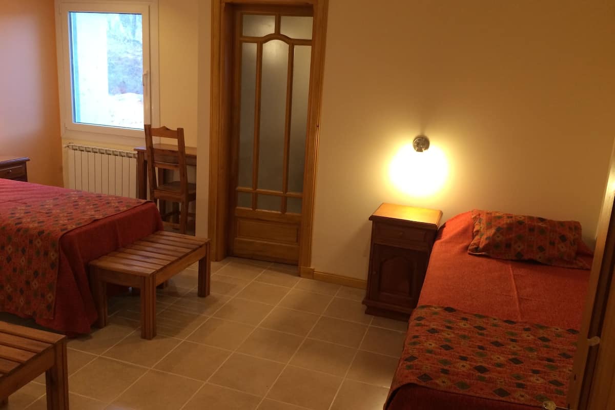 A room with two beds, a chair, a table, a window, a door, and a wall-mounted light.