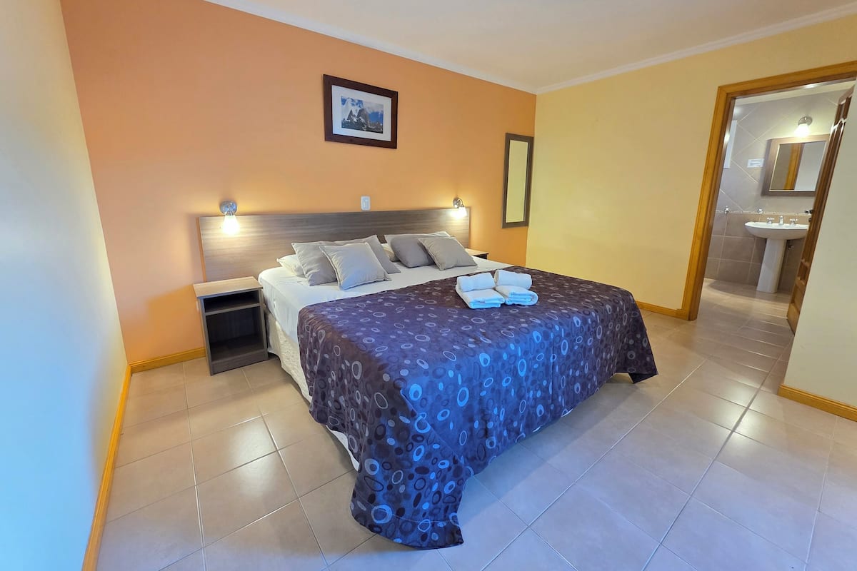 A hotel room with a bed, bedside tables, a framed picture on the wall, and a bathroom visible through an open door.