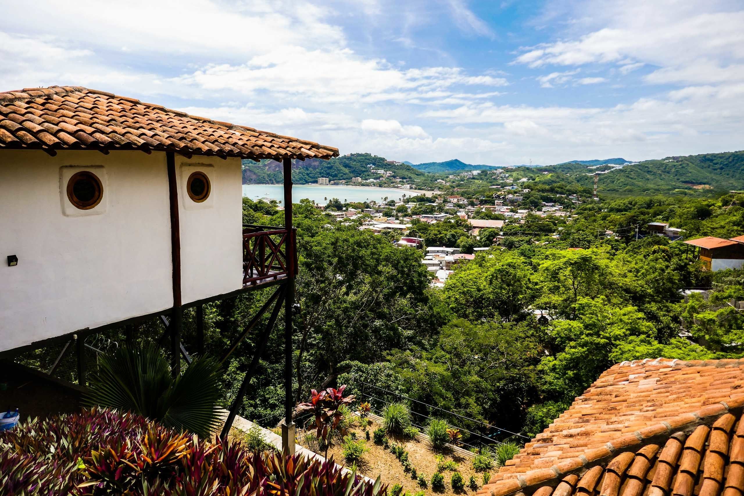 Un edificio blanco con tejado de tejas, un balcón y vistas a un pueblo costero y colinas.
