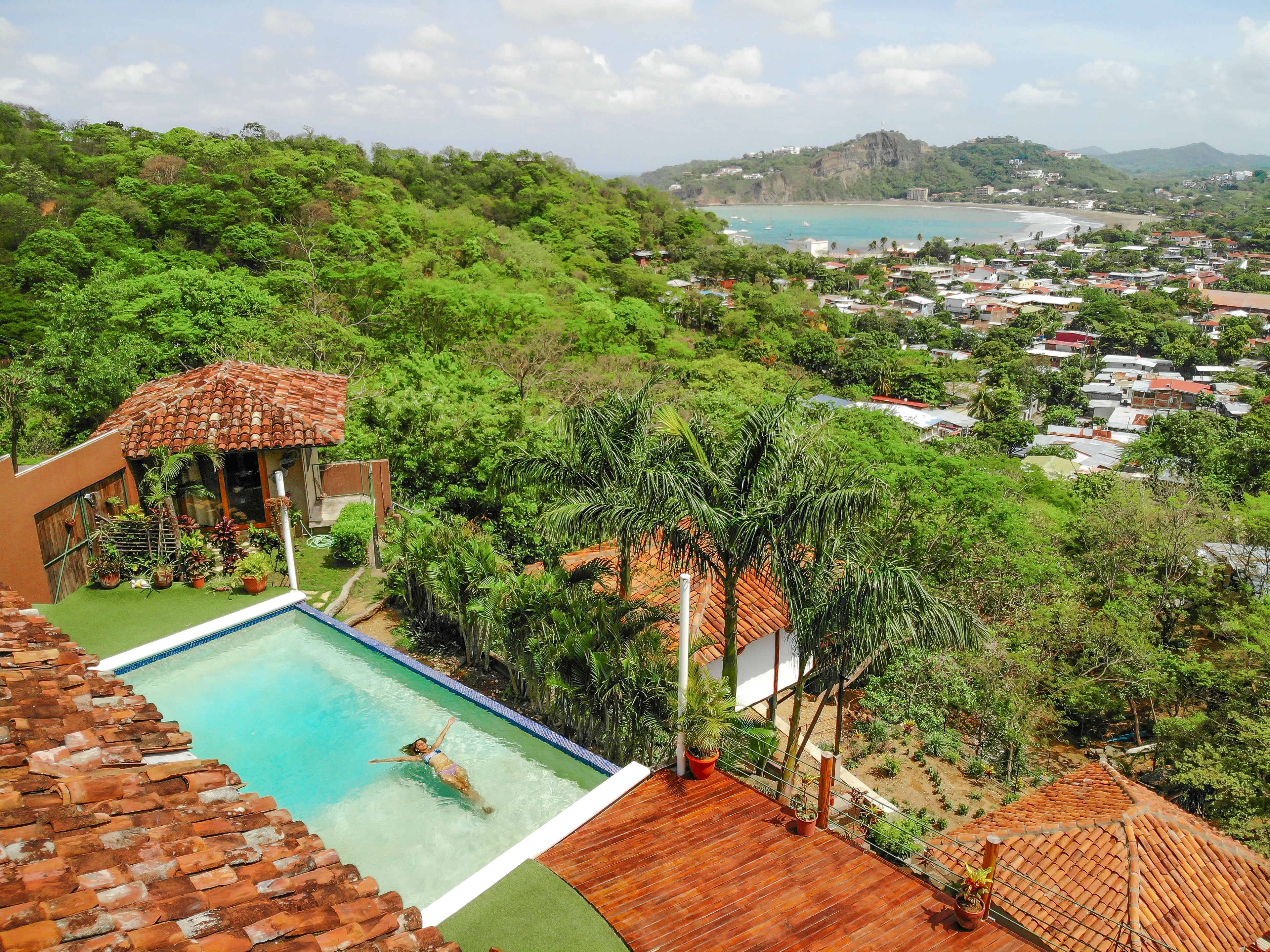 A swimming pool with a person swimming, surrounded by lush greenery and a view of a coastal town.