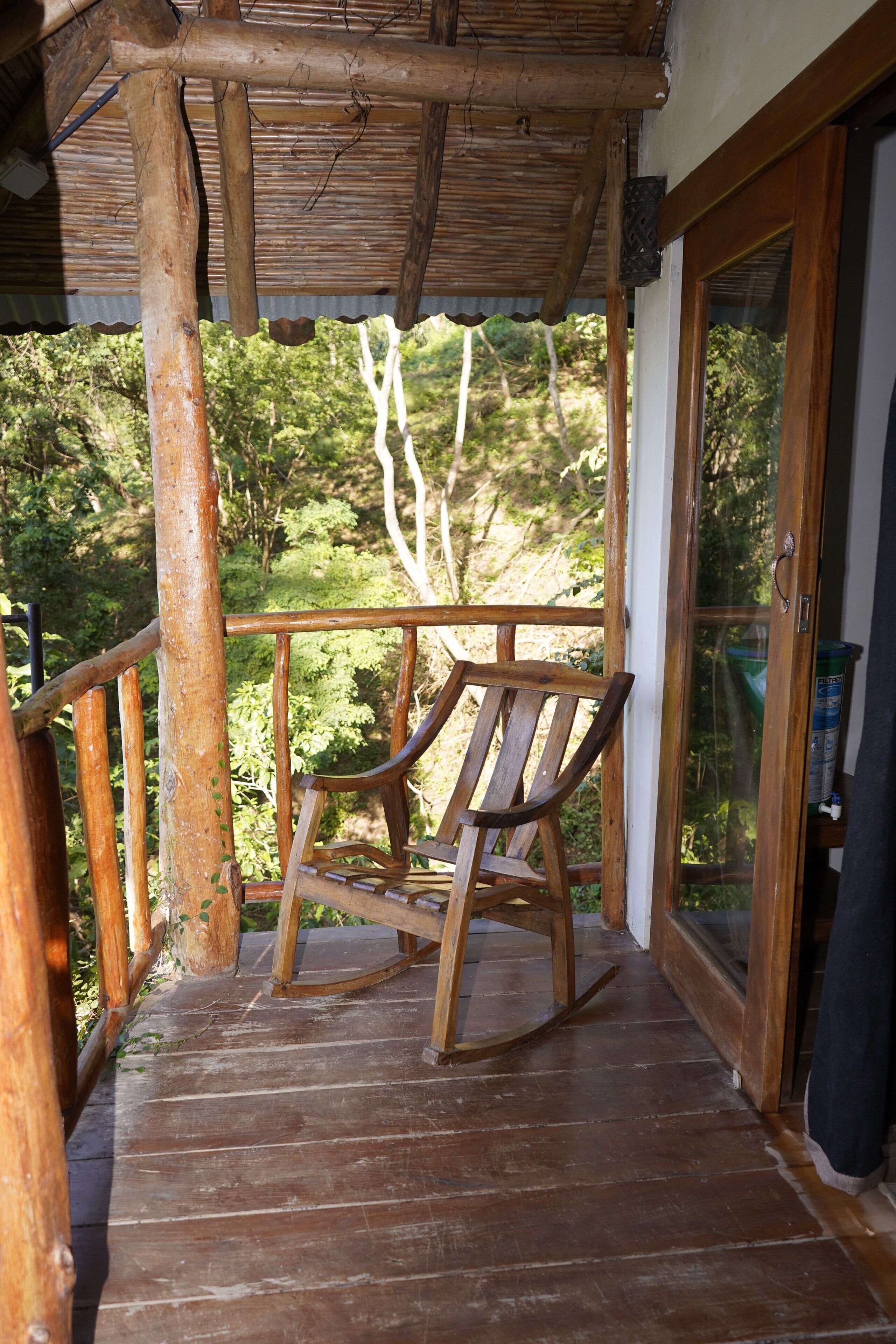 Una terraza de madera con una mecedora, un ventanal de vidrio y vistas al verdor.