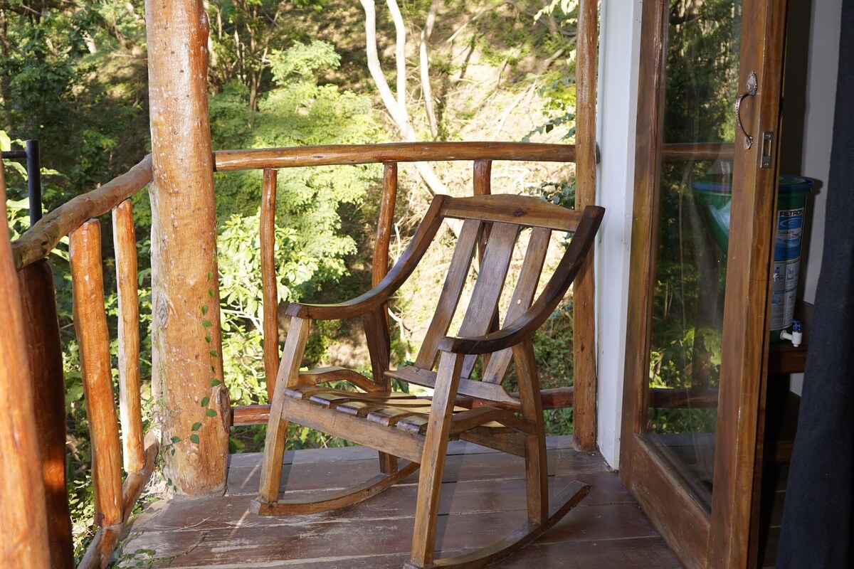 Une terrasse en bois avec un fauteuil à bascule, une porte vitrée et une vue sur la verdure.