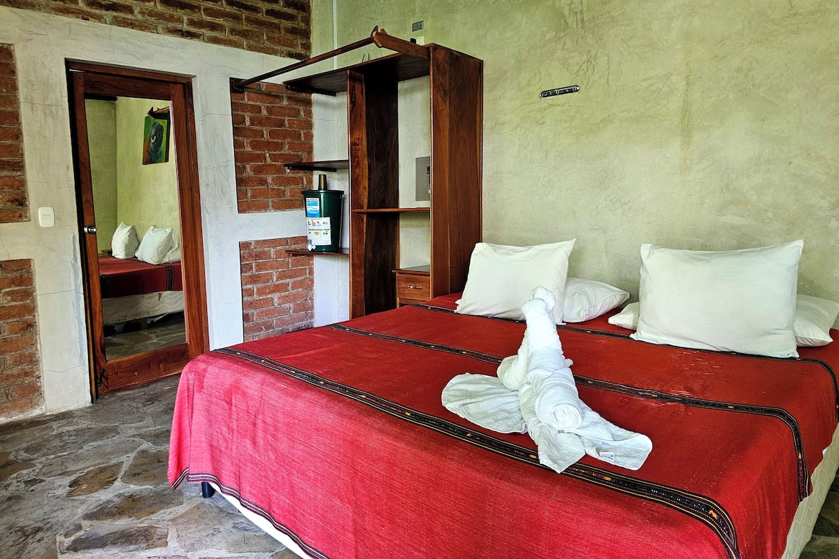 Een bed met een rode sprei en witte kussens, een houten hoofdbord, een bakstenen muur en een klein groen apparaat op een plank.