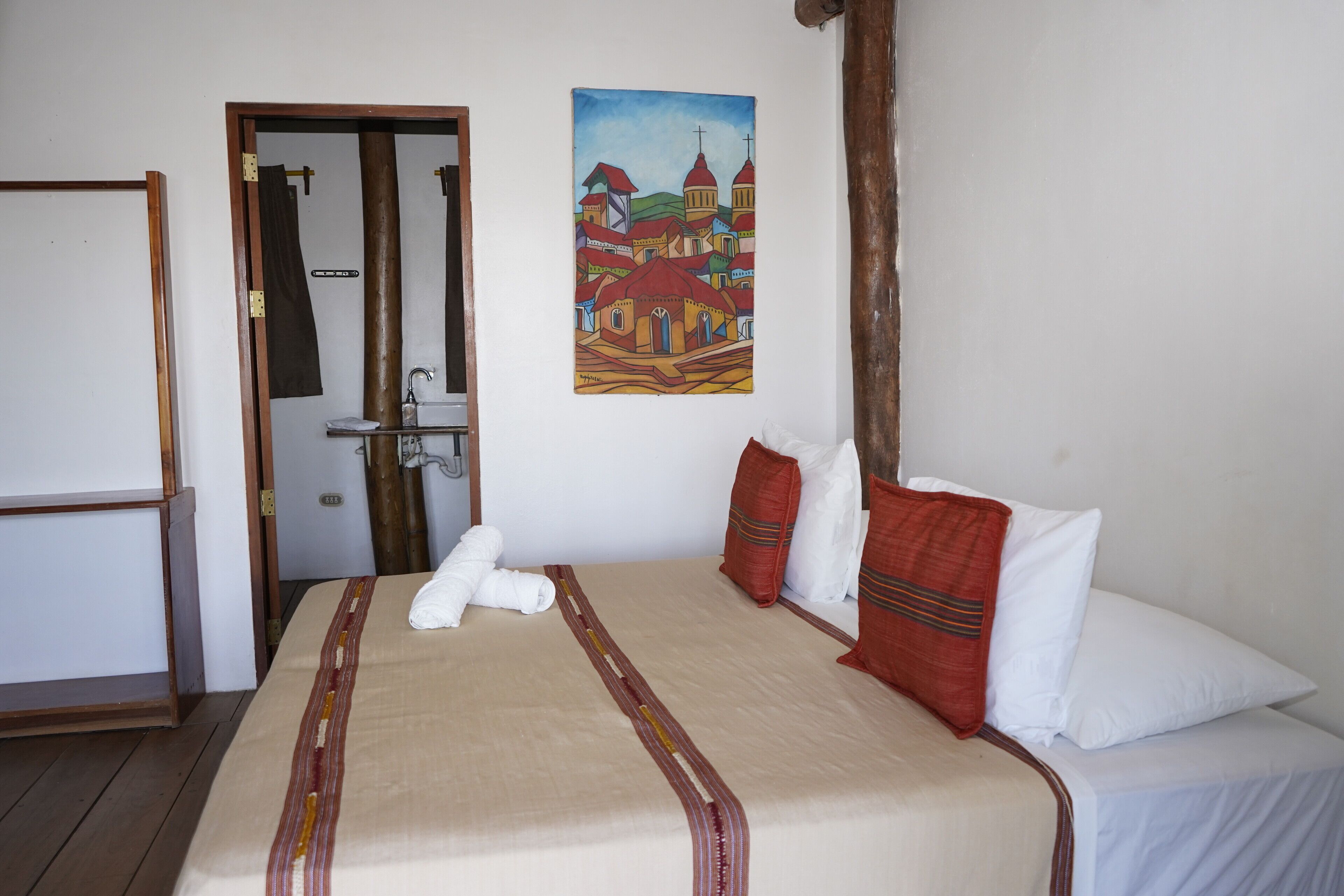 A hotel room with a bed, a painting of a town, and a door leading to a bathroom.