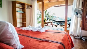 Een slaapkamer met een bed, een houten plank, een ventilator en uitzicht op een hangmat en buitenruimte.